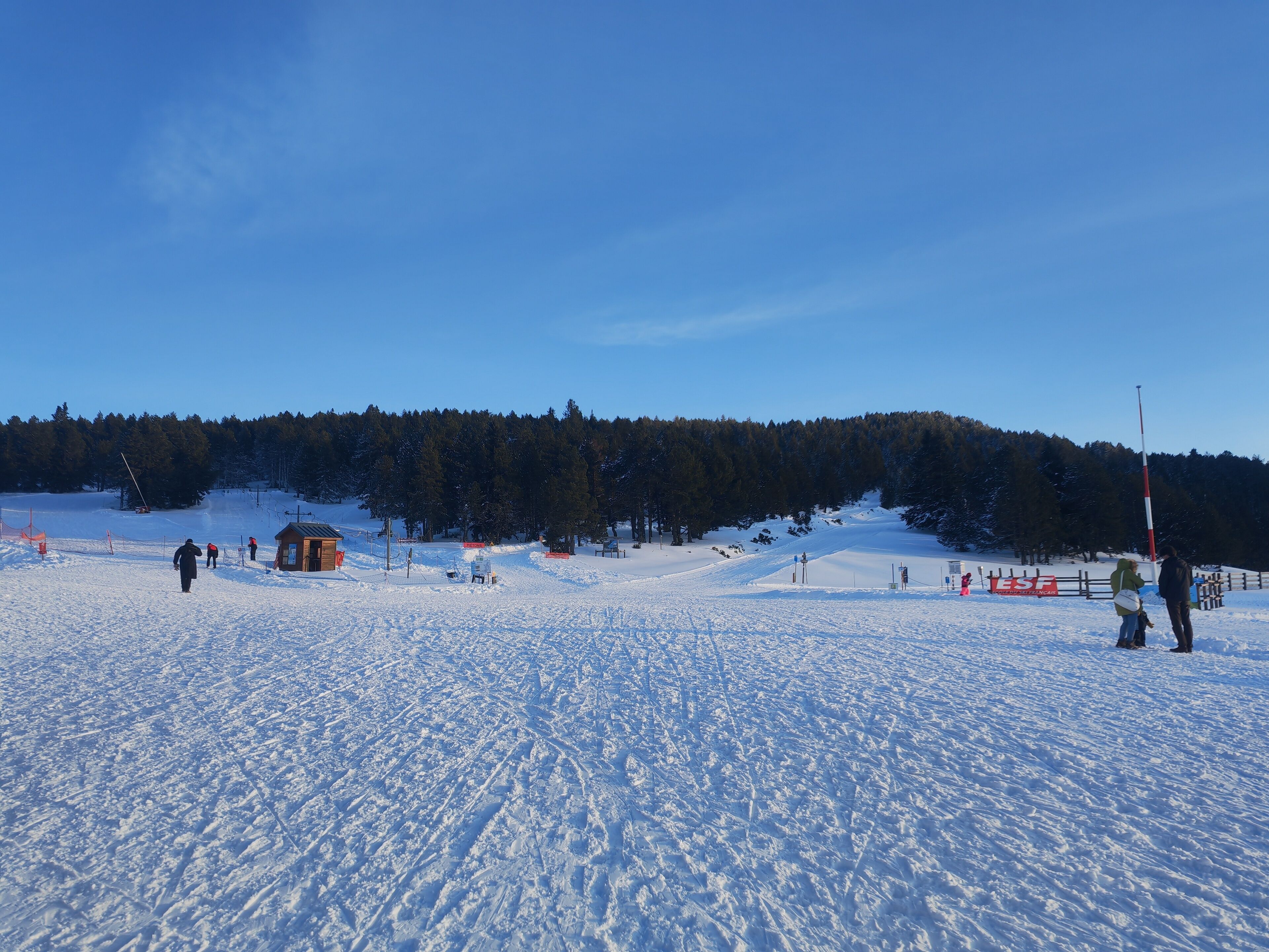 Pista d'esquí nevada amb una cabana de fusta, un teleàs i un turó forestal al fons.