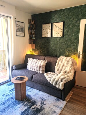 Ein modernes Wohnzimmer mit einem grauen Sofa, zwei flauschigen Decken, einem kleinen Holznachttisch mit einem Notizbuch und einem gemusterten Teppich.