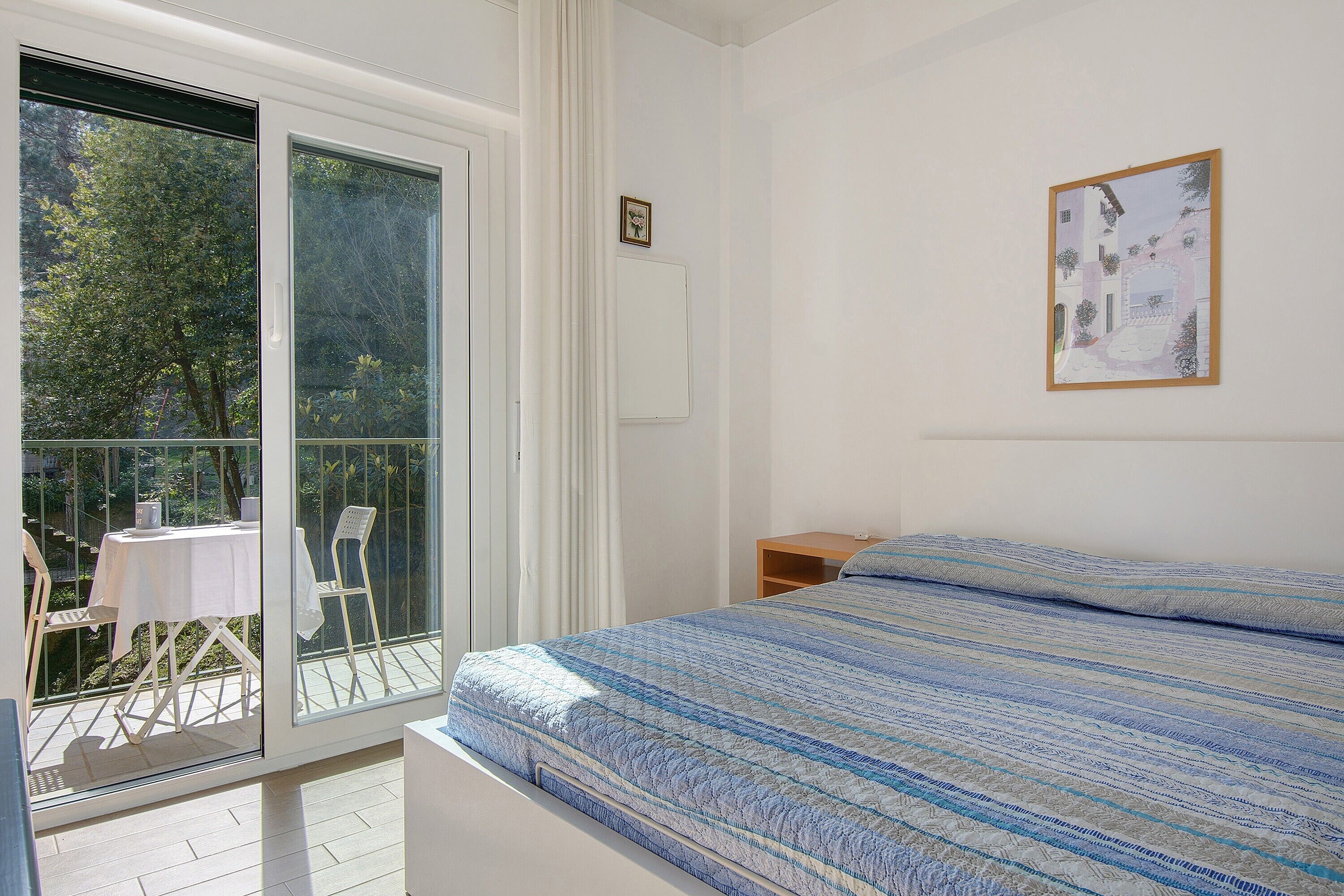 A bedroom with a bed, a window, a balcony with a table and chairs, and a framed picture on the wall.