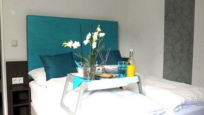 A neatly made bed with a teal headboard, a bedside table with a lamp, a tray with a bottle of orange juice, a glass of water, and a potted plant with white flowers. - Koblenz Valley Hotel Budget Double Room (Bendorf)