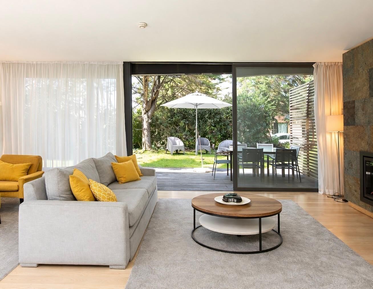 A modern living room with a grey sofa, yellow cushions, a round coffee table, and a large sliding glass door leading to an outdoor dining area.