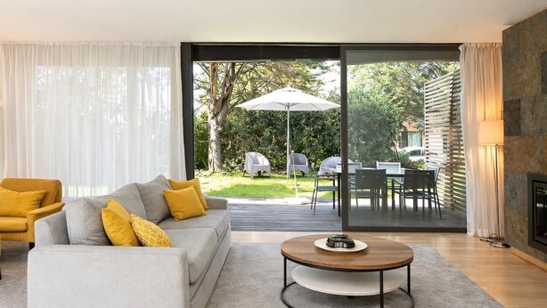 Un salon moderne avec un canapé gris, des coussins jaunes, une table basse ronde et une grande porte-fenêtre coulissante donnant sur un espace repas extérieur.