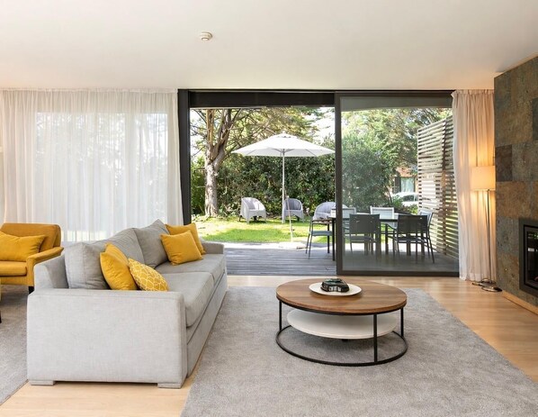 A modern living room with a grey sofa, yellow cushions, a round coffee table, and a large sliding glass door leading to an outdoor dining area. - Troia by the Ocean (Setúbal)