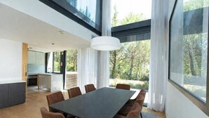 Un espace repas moderne, doté d’une grande table et de chaises, d’un luminaire suspendu et de grandes fenêtres offrant une vue sur l’extérieur.