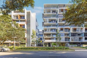 Un moderno edificio residencial de varios pisos con balcones, una estructura vertical central y un jardín bien cuidado.