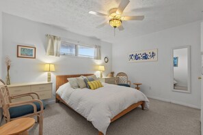 Ein Schlafzimmer mit einem Bett, zwei Nachttischen, einem Sessel, einem Schreibtisch, einem Deckenventilator und einem Fenster mit Vorhängen.