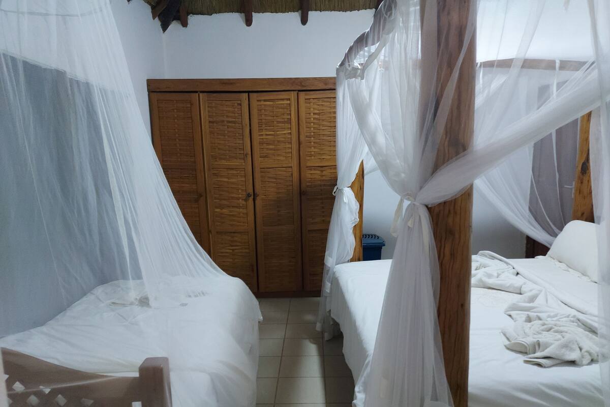 Une chambre avec deux lits, chacun recouvert de draps blancs et surmonté d’un dais. On aperçoit, au fond, une armoire en bois et une poubelle bleue.