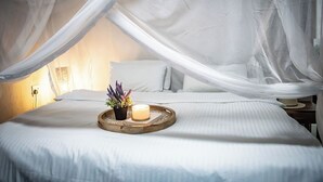 Una cama bien tendida con una colcha blanca, una mesita de noche con una vela y una planta, y un dosel sobre la cama.