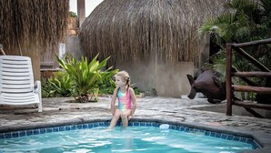Niño sentado en una pileta pequeña con techo de paja y cerca de madera al fondo.