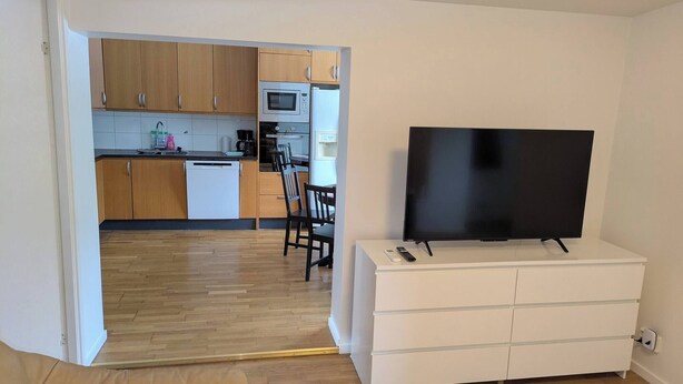 Een moderne woonkamer met een flatscreen televisie op een wit kastje, houten vloeren en een keuken op de achtergrond.