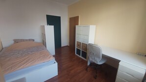 A bedroom with a bed, a desk, and a chair. There is a wardrobe with multiple drawers and a door.