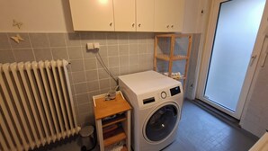 A laundry room with a front-loading washing machine, a radiator, a small wooden shelf, and a door to the outside.