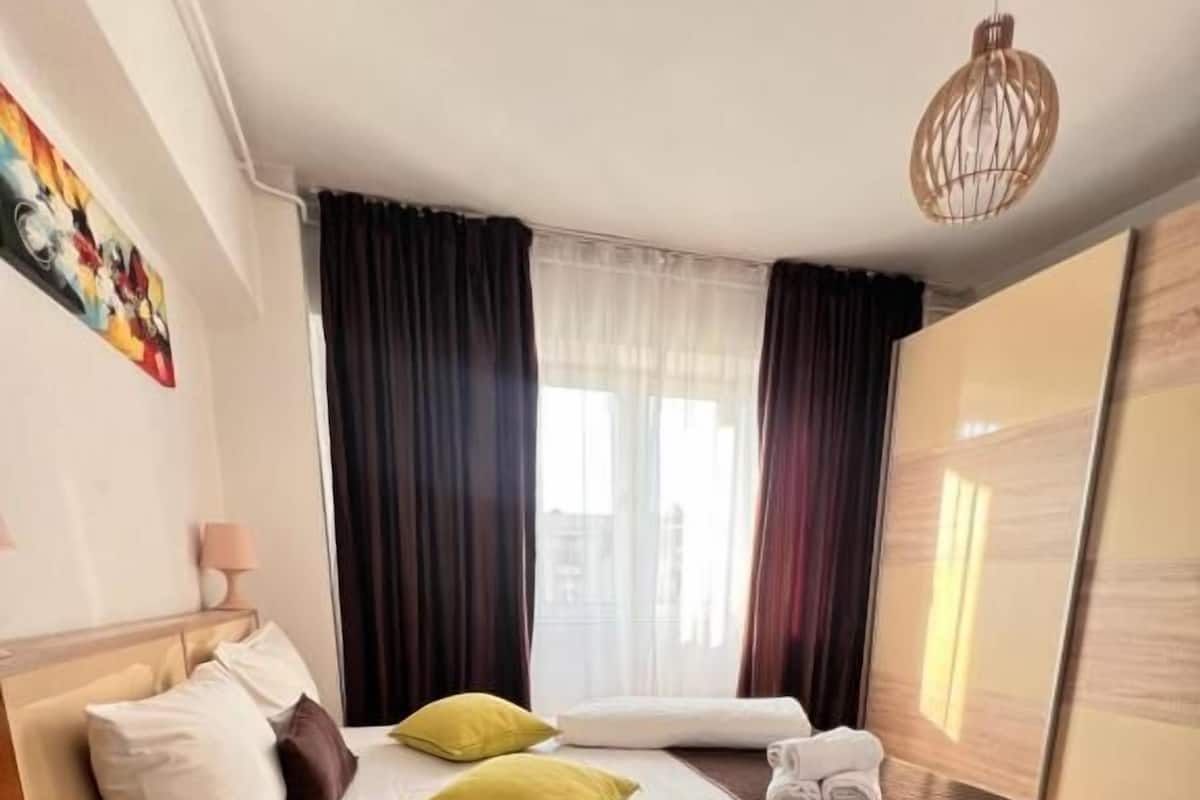 Un dormitorio moderno con cama, almohadas, una mesita de noche, lámpara, alfombra estampada y vista a la ciudad por la ventana.