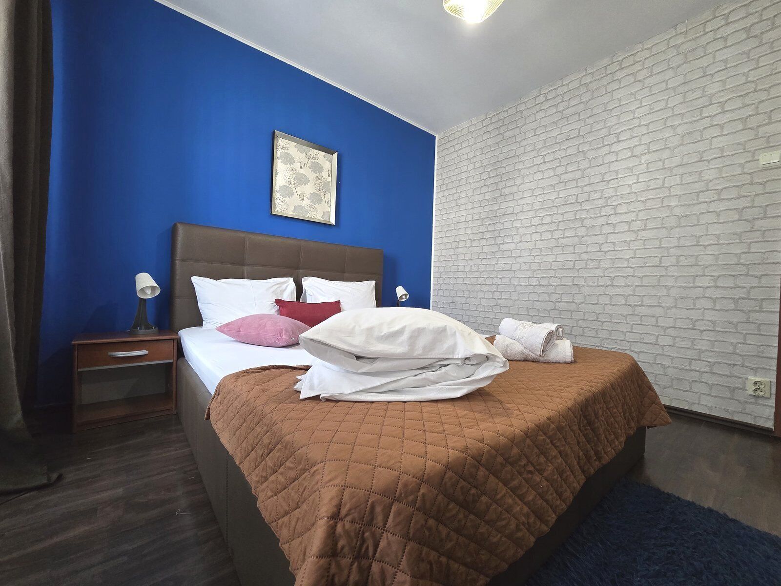 Una camera d'albergo con una parete blu, una parete in mattoni bianchi, una coperta letto trapuntata marrone e un letto singolo con cuscini bianchi e rosa.