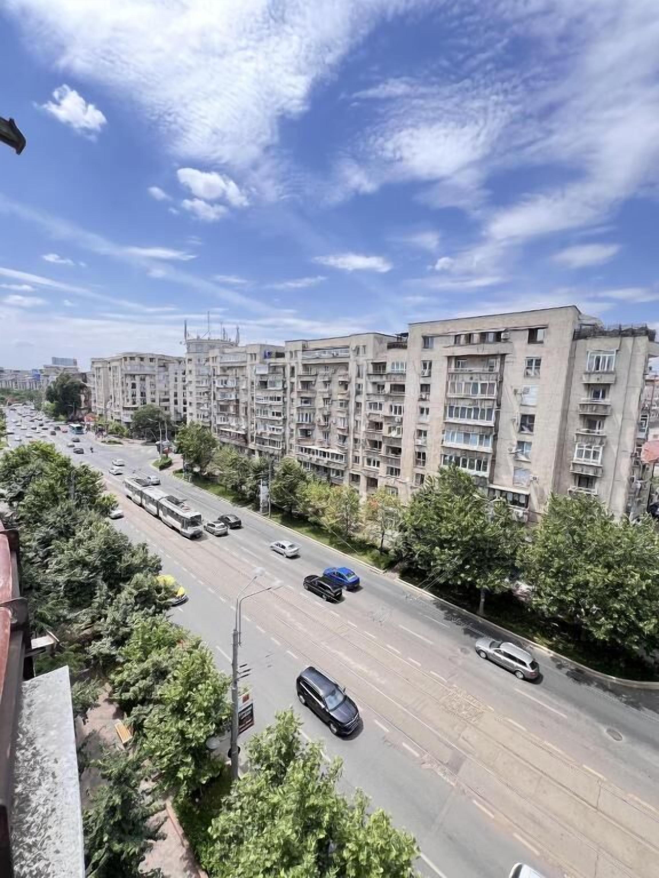 Un immeuble résidentiel à plusieurs étages, situé devant une large rue bordée d’arbres et de voitures garées.
