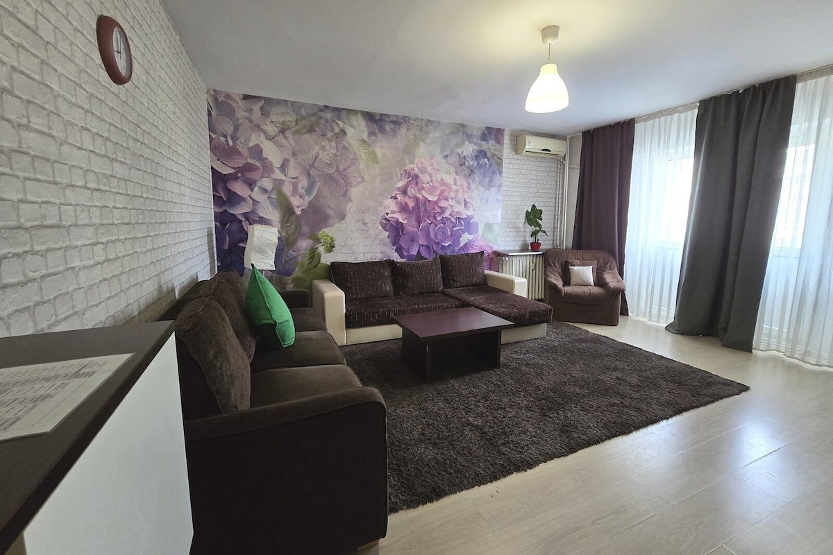 Una sala de estar moderna con una pared de ladrillo blanco, un mural floral, un sofá marrón, un cojín verde, una mesa de centro, una mesita auxiliar con una planta y una ventana con cortinas.