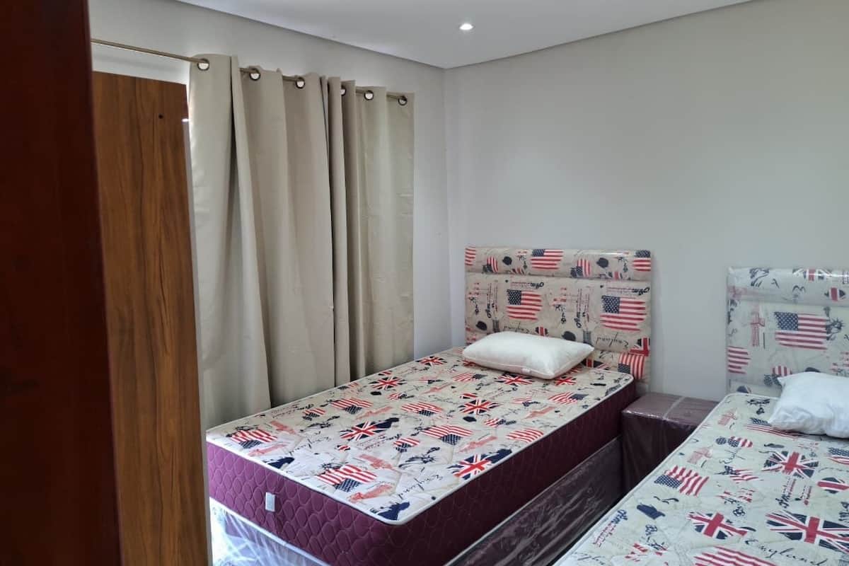 Une chambre à coucher avec deux lits, dont l’un est doté d’une tête de lit ornée d’un motif représentant un drapeau, et une fenêtre avec des rideaux.