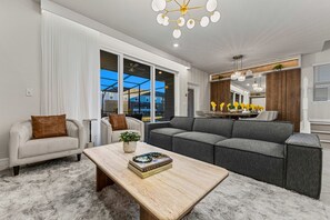A modern living room with a sectional sofa, a coffee table, and a dining area with a chandelier. - 5BR w Pool Grill Game Lounge Indoor Slide 16165 (Clermont)