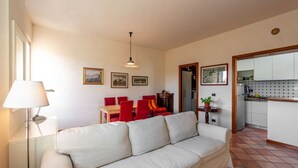 Sala de estar com sofá, mesa de jantar com cadeiras, luminária e área de cozinha visível por uma porta aberta.