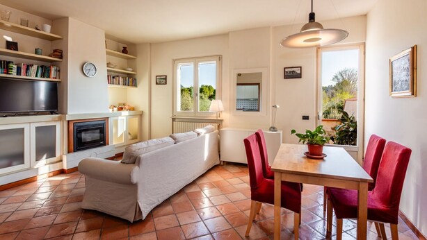 Uma sala de estar acolhedora com sofá, uma mesa de jantar com cadeiras, televisão e estantes com livros e objetos decorativos.
