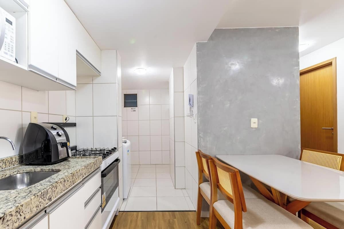 Uma cozinha compacta com micro-ondas embutido, torradeira e pia. Uma pequena área de refeições com mesa e cadeiras fica adjacente à cozinha.