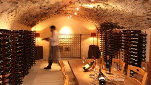 A wine cellar with racks of wine bottles, a table with wine glasses, and a person in the background. - Domaine les Mejeonnes (Valaurie)