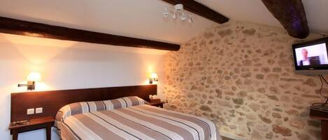 A hotel room with a stone wall, a bed with striped bedding, two bedside lamps, and a television on the wall.