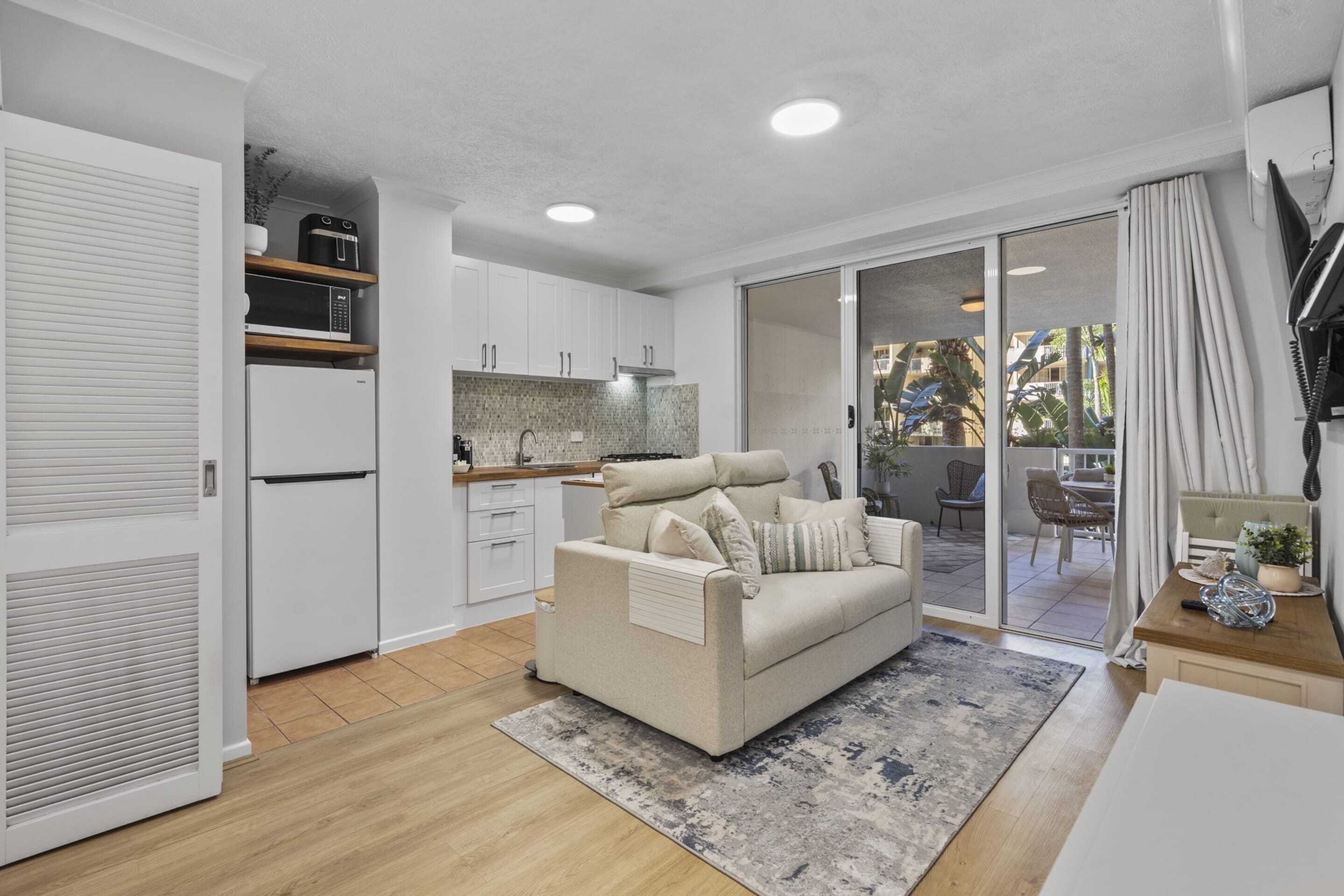 Una sala de estar moderna con un sofá, una mesa de centro y una zona de cocina con armarios blancos y un microondas.