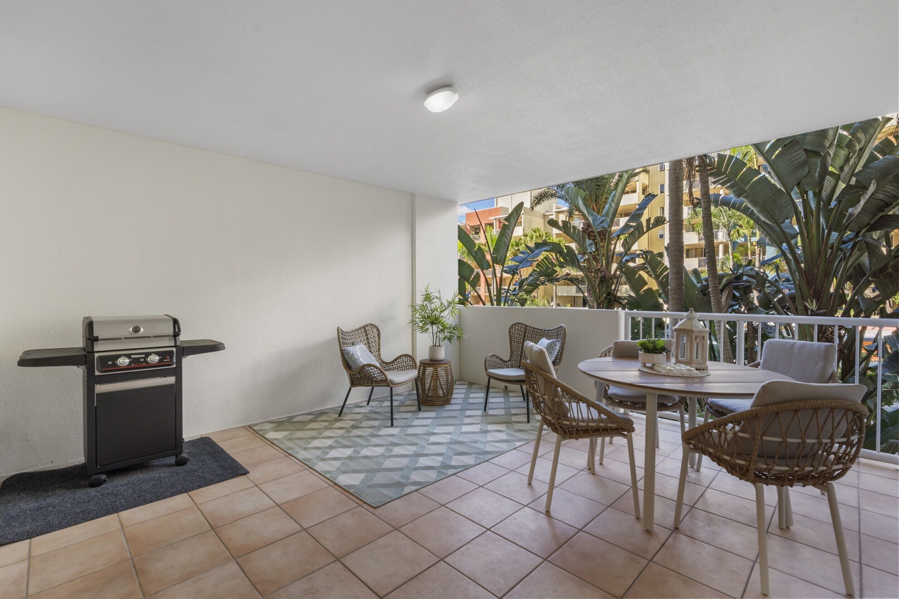 A balcony with a grill, wicker chairs, a small table, and a view of buildings and plants.