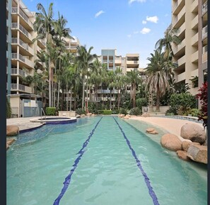 Une piscine à l’eau claire, entourée de palmiers et bordée d’un immeuble à plusieurs étages doté de balcons.