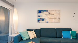 A modern living room with a dark blue sofa, a white side table, and a floor lamp. There is a large abstract painting on the wall and a window with a view of the outside. - Cozy Maspalomas Bungalow With Pool (San Bartolomé de Tirajana)