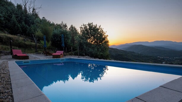 Une piscine rectangulaire à l’eau d’un bleu limpide, bordée d’une murette en pierre. On y trouve deux chaises longues rouges et un parasol bleu, près de la piscine. En arrière-plan, on aperçoit une colline boisée et un coucher de soleil sur des montagnes lointaines.