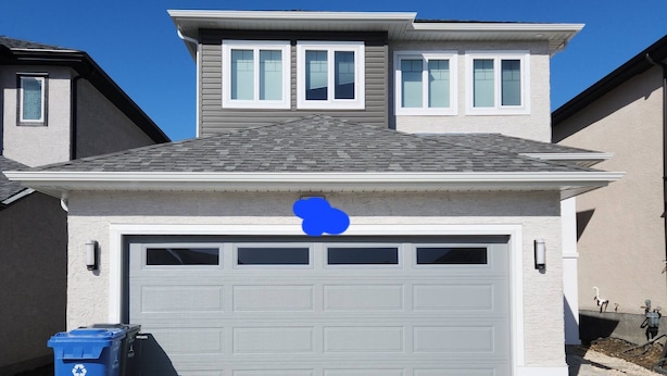 Una casa de dos pisos con un garaje, un contenedor azul para reciclaje y un cielo azul despejado.