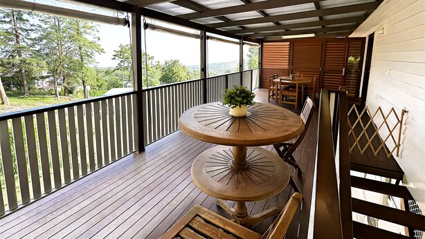Een overdekt terras buiten met een houten tafel en stoelen, uitzicht op de omliggende groene omgeving, en een houten balustrade.