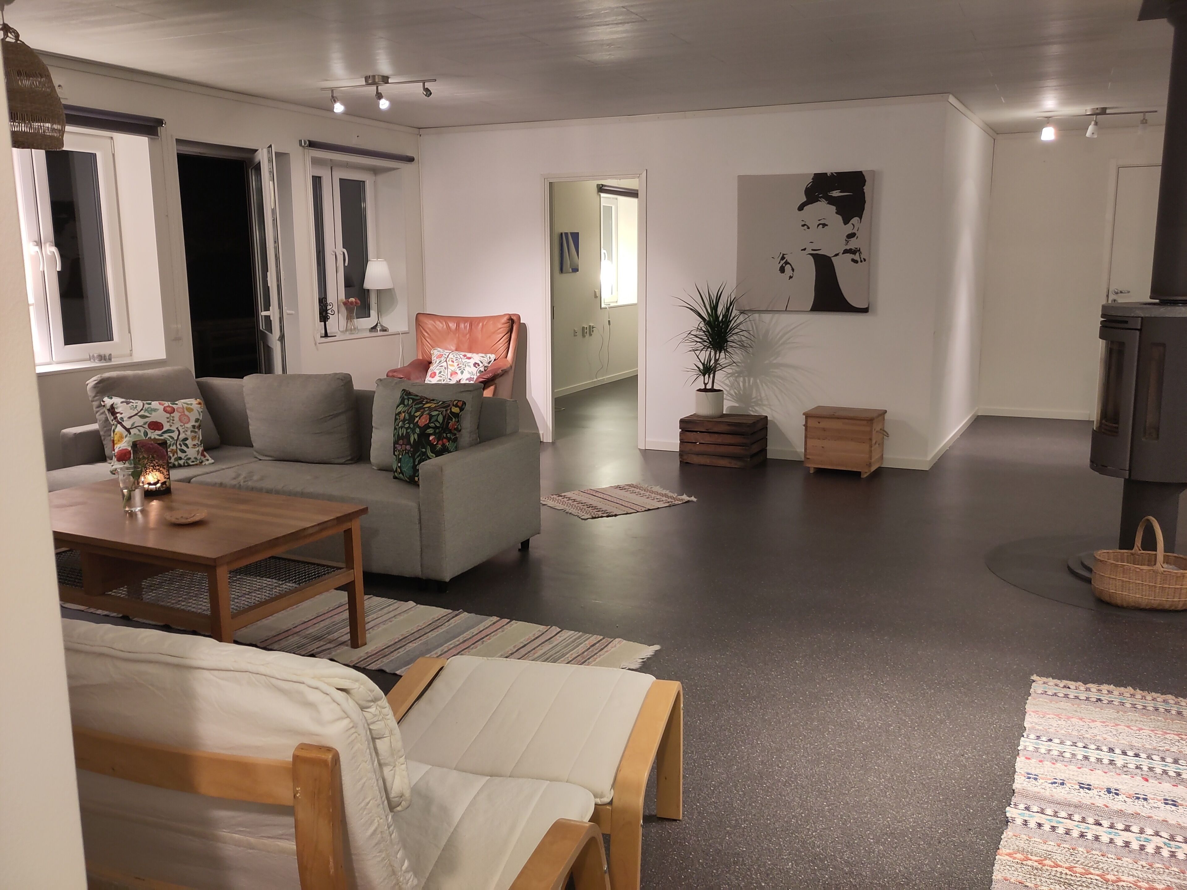 A modern living room with a sofa, a wooden coffee table, a small table with a candle, and a chair. There is a potted plant and a small wooden chest in the corner.
