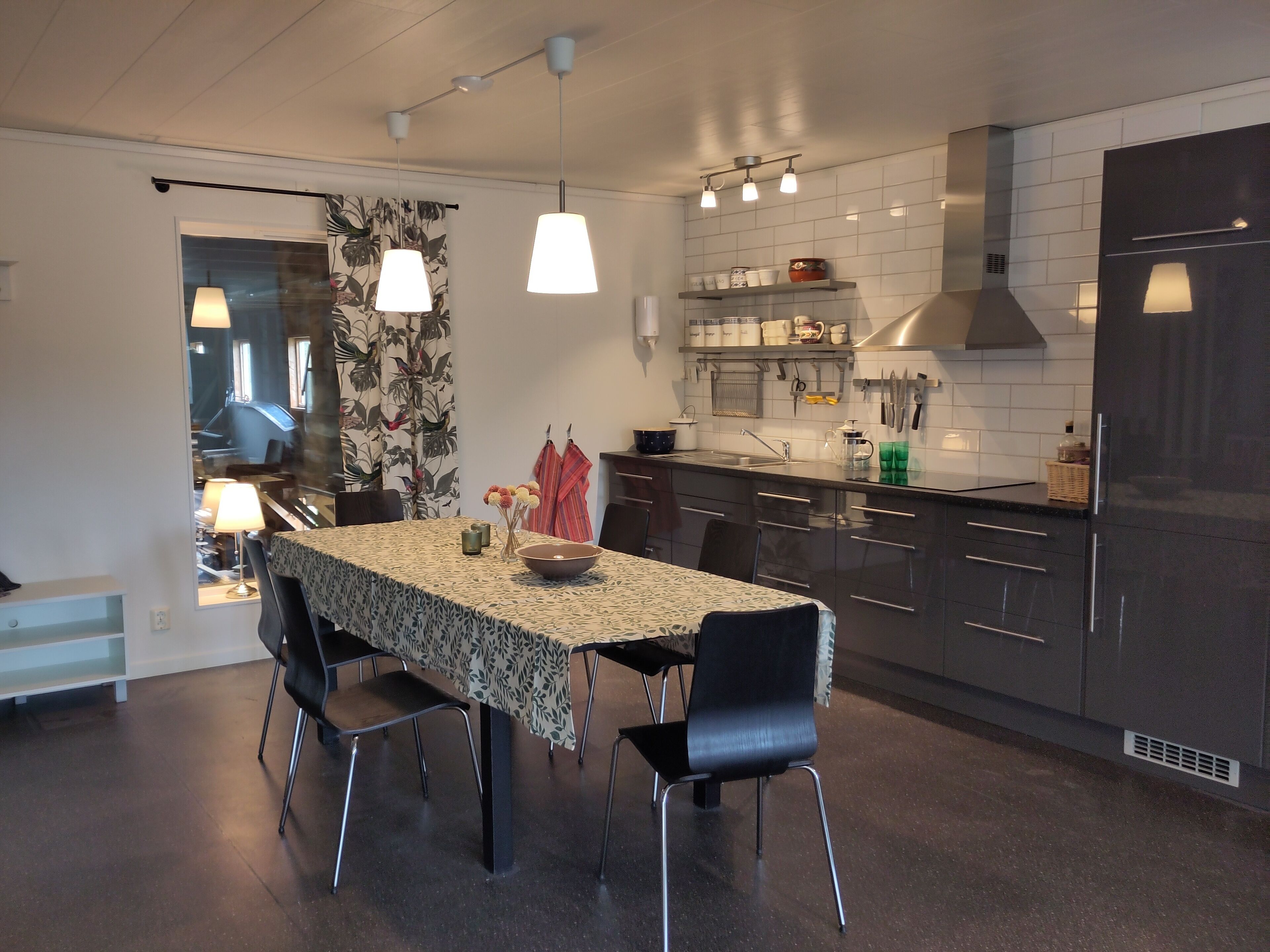 A modern kitchen with dark cabinets, a dining table with chairs, pendant lights, and a view of the dining area through a glass door.