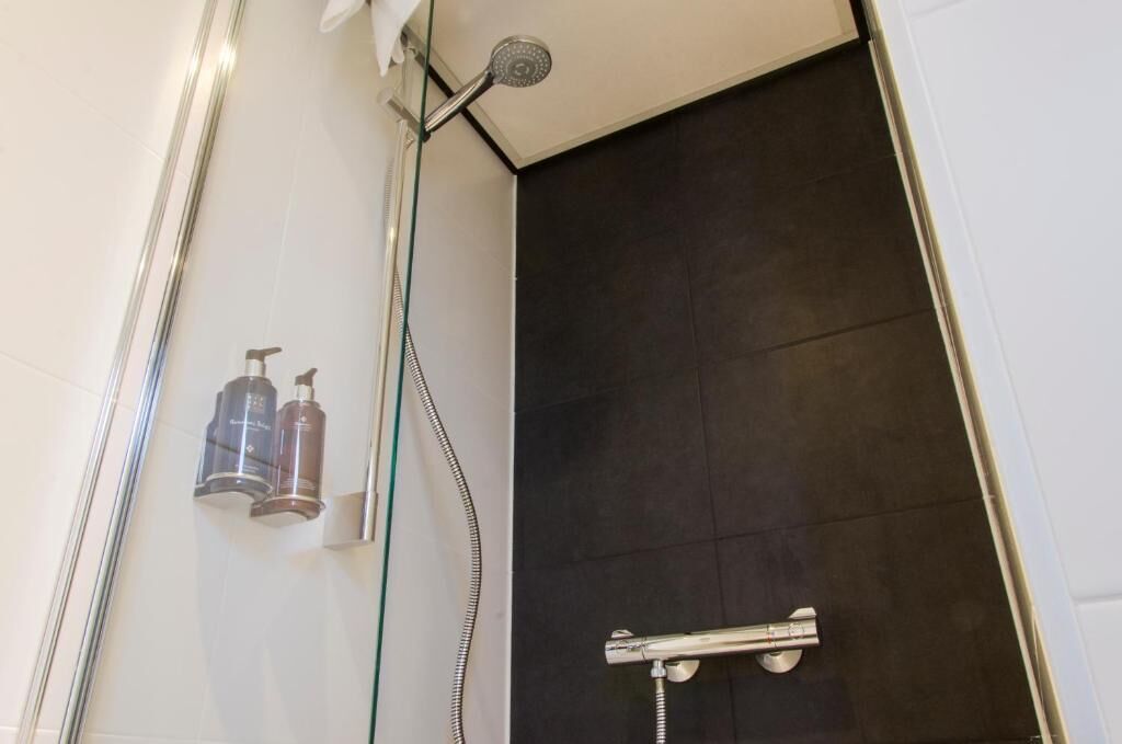 Un espace douche avec un mur recouvert de carreaux foncés, une paroi de douche en verre et deux bouteilles de shampoing.