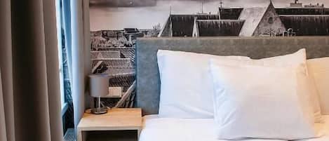 A hotel room with a bed, a nightstand, a lamp, and a view of buildings through the window.