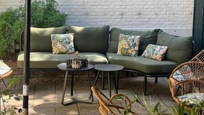 Ein grünes Sofa mit floralen Kissen, ein kleiner runder Tisch und Korbstühle auf einer Terrassenanlage. Im Hintergrund eine weiße Ziegelwand.