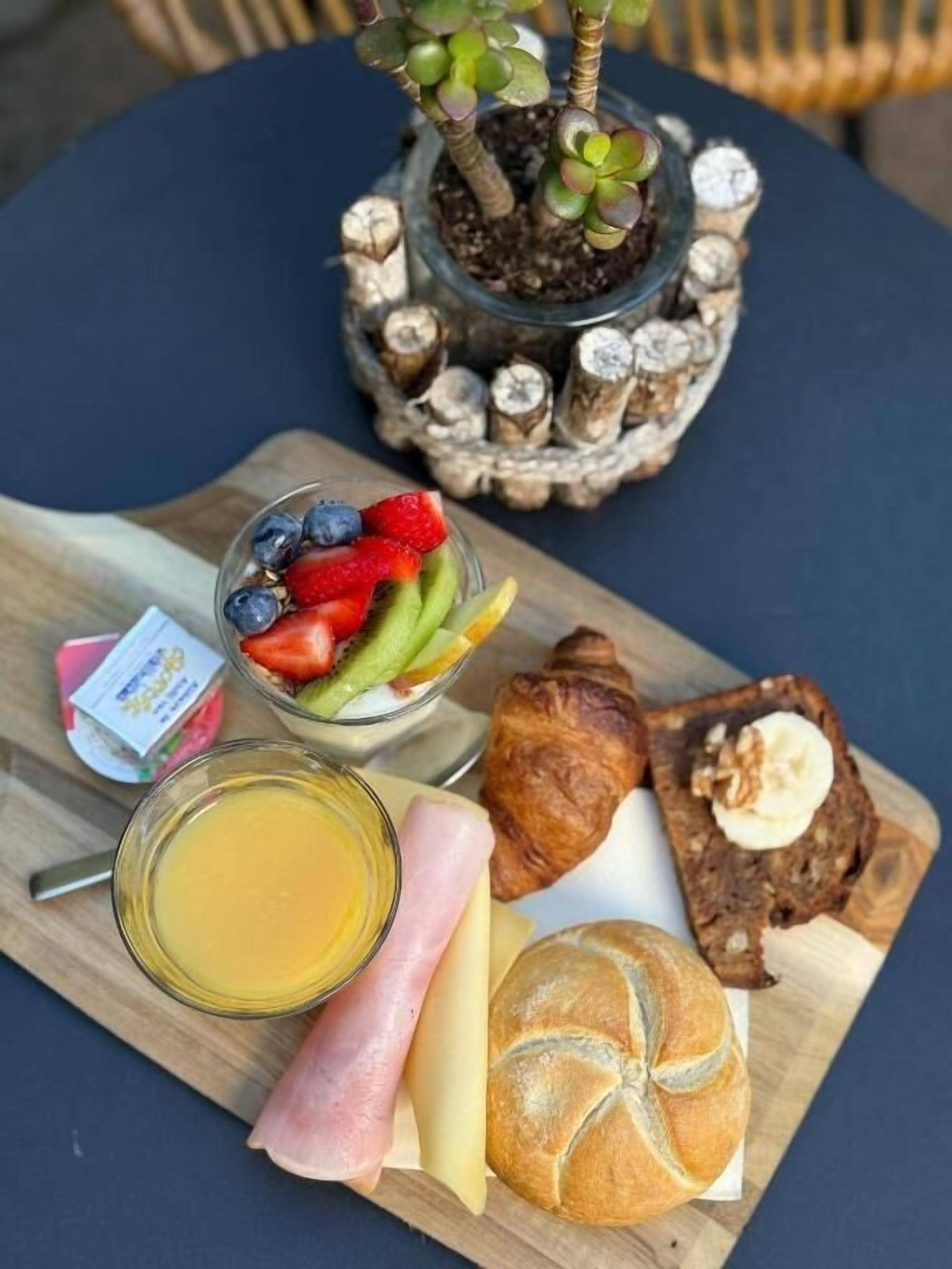 Ein Frühstückstisch mit verschiedenen Speisen, darunter Früchte, Brot und einer Pflanze im Glas.