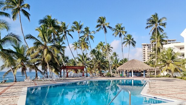 Uma área de piscina com espreguiçadeiras, uma estrutura com telhado de palha e palmeiras.