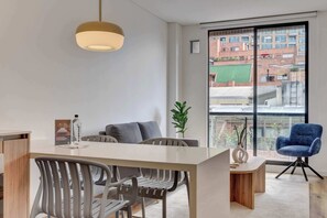 Une pièce moderne et bien éclairée, comprenant un coin repas, une grande fenêtre donnant sur des immeubles et une chaise bleue.