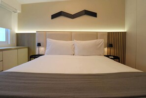 Uma cama bem arrumada com cabeceira, duas mesas de cabeceira com abajures e uma janela com persianas.