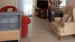 Ein Wohnzimmer mit Fernseher, einem roten Sofa, einem beigen Sessel und einer Essecke mit Tisch und Stühlen.