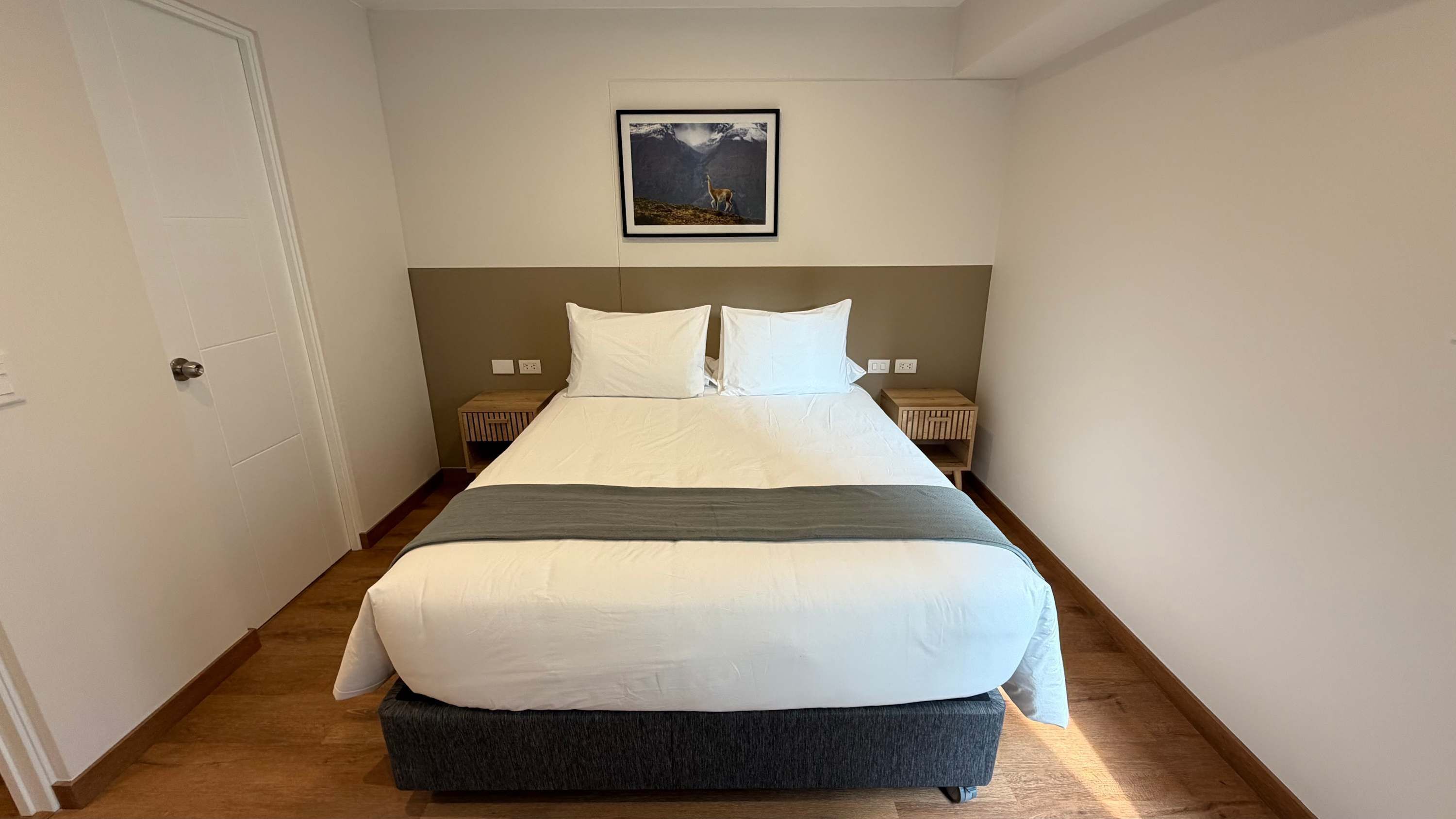 Una cama bien hecha con ropa de cama blanca y gris, dos mesitas de noche con lámparas, un cuadro en la pared y suelo de madera.