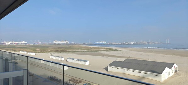 Vista de uma varanda com vista para uma praia, com alguns edifícios, incluindo uma construção branca com telhado escuro.