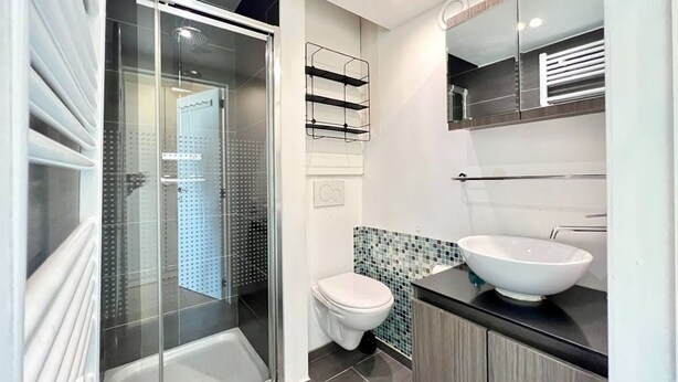 Ein modernes Badezimmer mit Duschkabine aus Glas, einer weißen Toilette, einer Rückwand aus Mosaikfliesen und einem großen, weißen Waschbecken auf einer schwarzen Arbeitsplatte.