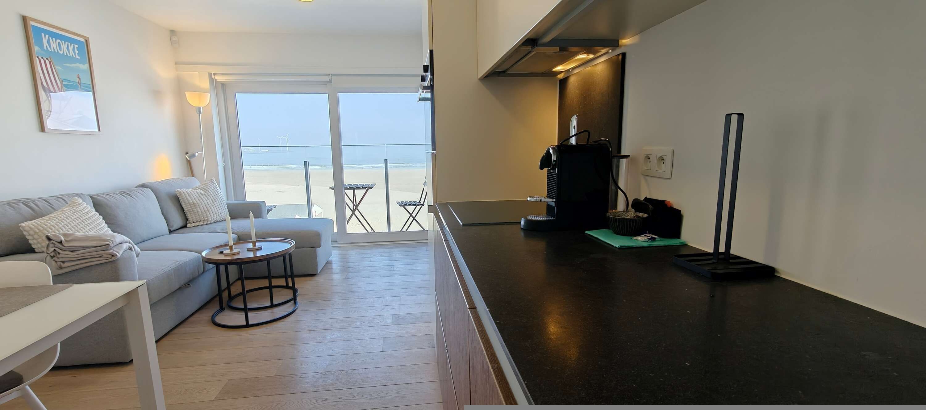 Una sala de estar moderna con un sofá, una pequeña mesa redonda y vistas a la playa a través de amplias ventanas.