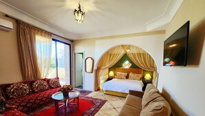 Una habitación de hotel con cama, sofá, televisor, candelabro y un amplio ventanal con cortinas.