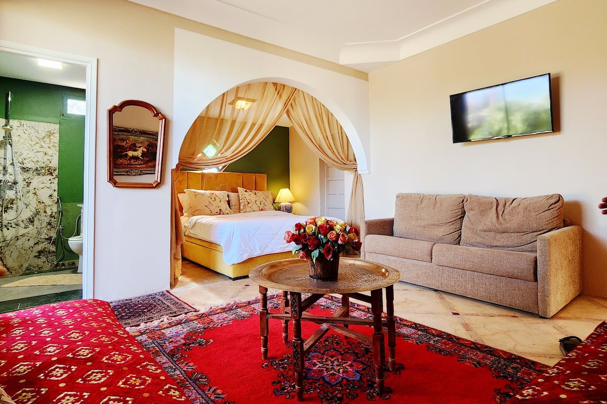 A room with a bed, a sofa, a small table with a vase, and a painting on the wall.